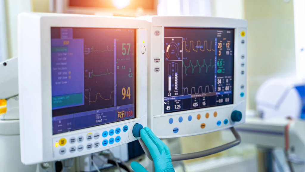 Monitor multiparamétrico em ambiente hospitalar exibindo sinais vitais como frequência cardíaca, pressão arterial e curvas respiratórias, com mão enluvadas ajustando o painel